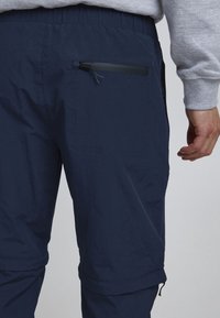 Pantalon léger bleu marine avec taille élastique, doté d'une poche arrière zippée et de fermetures éclair aux chevilles pour un ajustement réglable. Texture lisse.