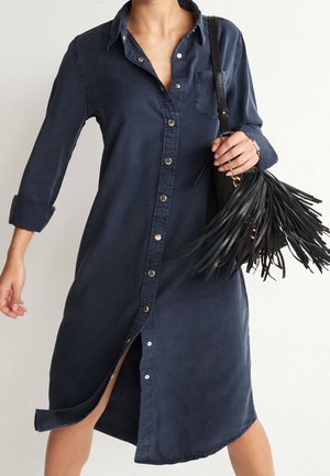 Robe bleu marine à boutons avec manches longues et poche poitrine. Présente une fente haute et une texture de tissu usé. Sac à franges noires en accent.