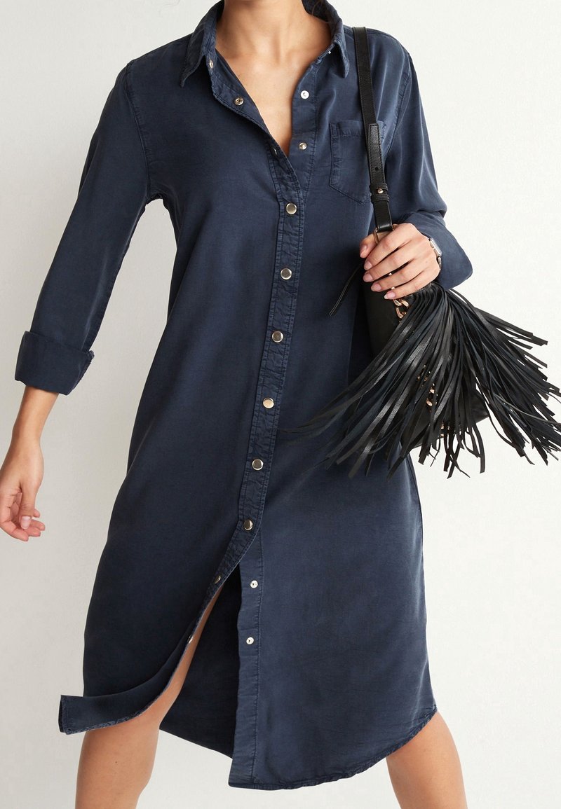Robe bleu marine à boutons avec manches longues et poche poitrine. Présente une fente haute et une texture de tissu usé. Sac à franges noires en accent.