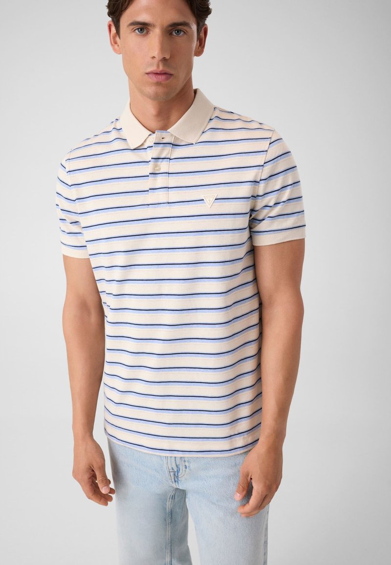 Jeune homme portant un polo beige à manches courtes avec des rayures horizontales bleues et un jean bleu clair, debout devant un fond uni.