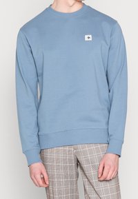 Jasnoniebieska bluza z miękkiego materiału, z okrągłym dekoltem i ściągaczami przy rękawach. Mała naszywka z logo na lewej piersi, minimalistyczny design.