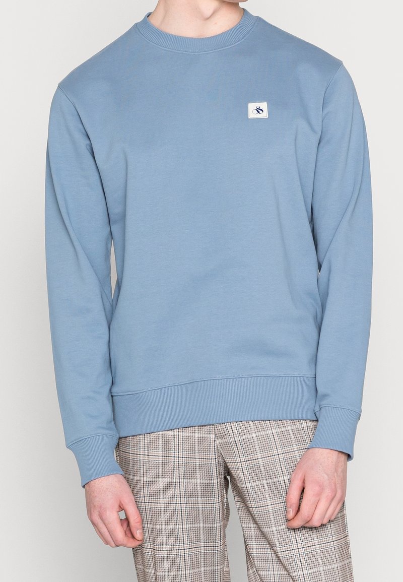 Jasnoniebieska bluza z miękkiego materiału, z okrągłym dekoltem i ściągaczami przy rękawach. Mała naszywka z logo na lewej piersi, minimalistyczny design.
