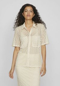 Camisa de botonadura en encaje color crema, con mangas cortas, dos bolsillos frontales y un patrón texturizado en toda la prenda. Ligera y semi-translúcida.