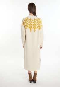 Beige neulemekko, jossa on pitkät hihat, olkapäillä lehtikuviollista keltaista brodeerausta, sivuhalkioita ja suora siluetti.