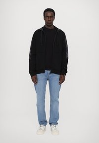 Crna hoodie s uzorkastim bočnim panelima, uparena s crnom majicom i svijetloplavim trapericama. Model nosi bijele tenisice, stoji naslonjen na bijelu pozadinu.