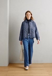 Gequilte marineblauwe jas met een kraag, knoopsluiting en lange mouwen, gecombineerd met lichtblauwe wijde jeans en een beige tas.