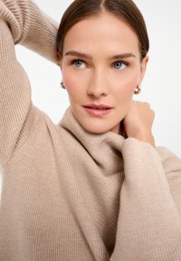 Beige ribbed knit sweater with a high collar. Soft texture, fitted sleeves, and relaxed draping around the neck. Model poses with one arm raised.