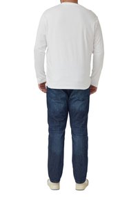 Weißes Langarmhemd kombiniert mit blauen Denim-Jeans. Das Hemd hat einen Rundhalsausschnitt; die Jeans sind leicht verjüngt und haben Gesäßtaschen. Die Schuhe sind hell.