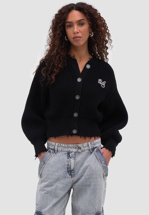 Kvinde med krøllet hår iført en sort cropped knaptrøje med logoer og lyseblå højtaljede jeans med hænderne i lommerne.