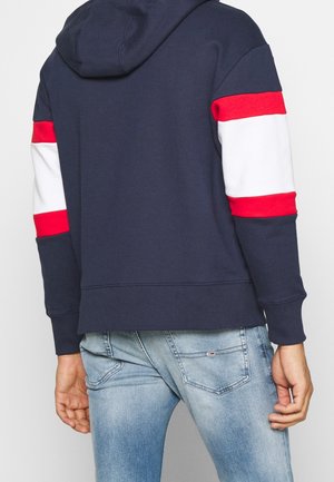 Osoba ubrana w granatową bluzę z kapturem z czerwonymi i białymi paskami na rękawach oraz jasnoniebieskie, wyblakłe dżinsy, widziana od tyłu.