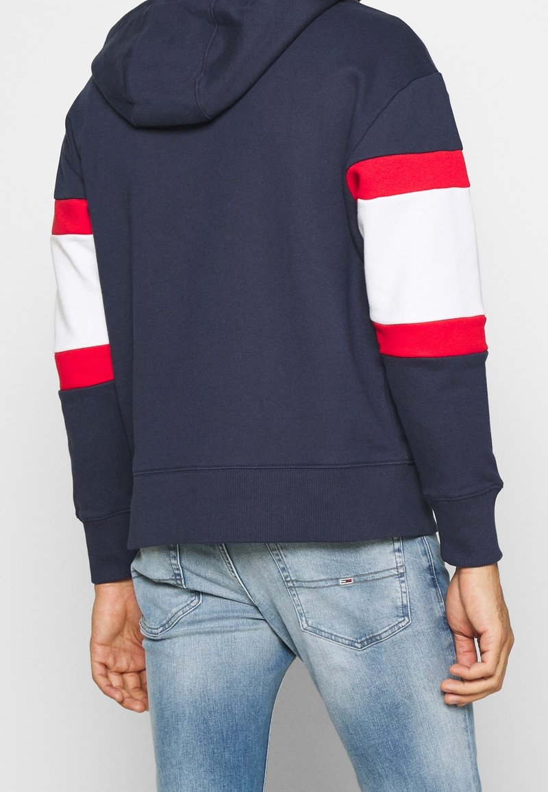Personne portant un sweat à capuche bleu marine avec des bandes rouges et blanches sur les manches et un jean délavé bleu clair, vue de dos.