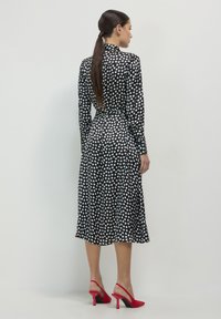 Schwarzes Kleid mit weißen Punkten, lange Ärmel, Taillengürtel, fließender Saum. Kombiniert mit roten Slingback-Absätzen. Glatte Satin-Textur.