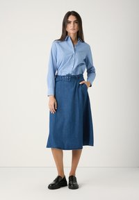 Frau in einem hellblauen Hemd mit Knöpfen, das in einen taillierten, knielangen Jeansrock gesteckt ist, mit schwarzen Slippern, steht vor einem schlichten weißen Hintergrund.