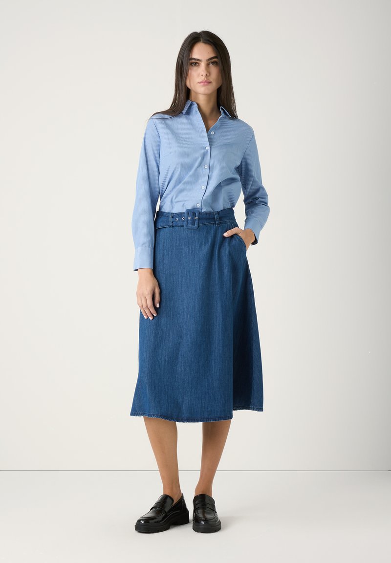 Frau in einem hellblauen Hemd mit Knöpfen, das in einen taillierten, knielangen Jeansrock gesteckt ist, mit schwarzen Slippern, steht vor einem schlichten weißen Hintergrund.