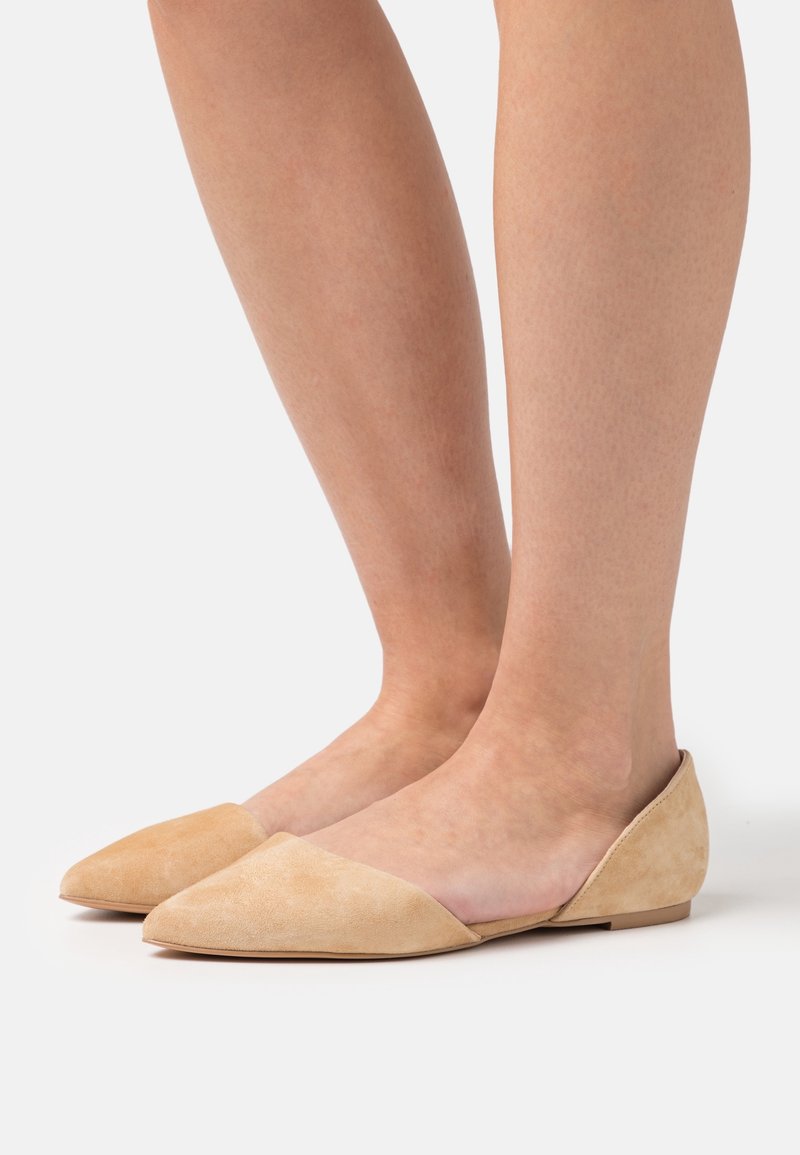 Chaussures plates en daim beige avec un bout pointu, un design profil bas, et un accent en daim plus foncé contrastant sur le bord avant.