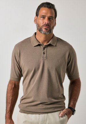 Mand med skæg iført brun kortærmet poloshirt og lyse bukser, står mod en ensfarvet lys baggrund med hånden i lommen.
