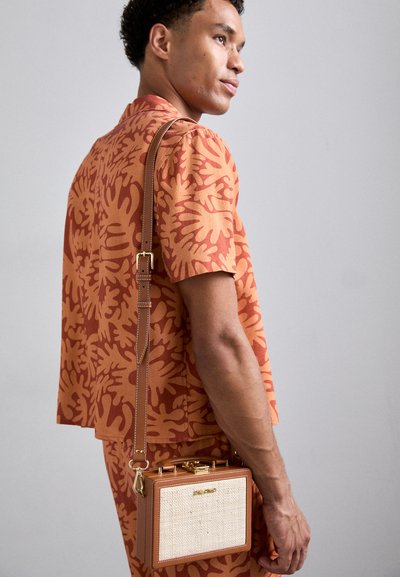 Brown handbag with gold hardware, rectangular shape, woven texture front, and a brown shoulder strap. Model wearing orange patterned outfit.