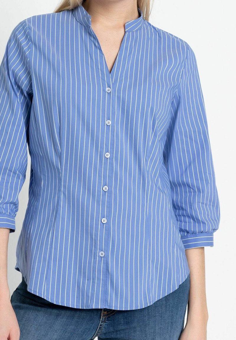 Femme portant une chemise bleue à boutons avec des rayures verticales blanches et des manches trois-quarts, associée à un jean bleu.