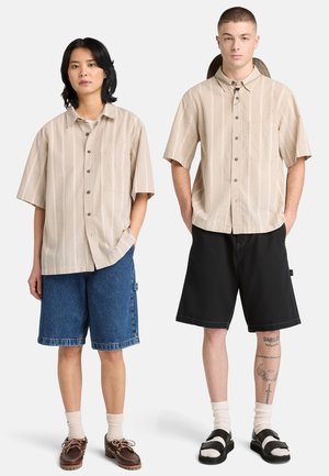 Due modelli stanno fianco a fianco indossando camicie beige a maniche corte con bottoni e tasche sul petto; uno indossa pantaloncini di jeans, l'altro pantaloncini neri.