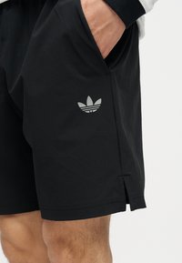 Person wearing black Adidas shorts with hand in pocket, showing Adidas trefoil logo on thigh.