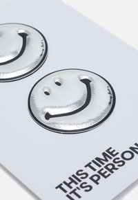 Dois autocolantes circulares prateados de sorrisos com contornos pretos num fundo branco, acompanhados de texto em negrito a dizer "Desta vez é pessoal."