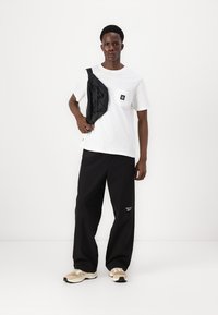 White cotton t-shirt with pocket, black loose-fit trousers, and beige sneakers. Black crossbody bag adds contrast and functionality.