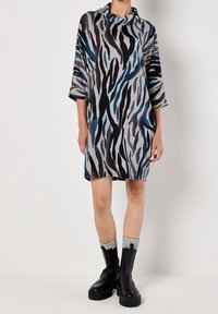 Robe grise et noire avec des rayures bleues, col bénitier et manches trois-quarts. Le matériau semble texturé, arrivant à mi-cuisse. Bottes compensées noires.