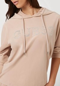Felpa rosa chiaro con la parola "GUESS" in strass argentati, con cappuccio regolabile e una texture di stoffa morbida.