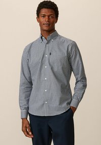 Chemise à carreaux bleue et blanche, à manches longues, avec col, en tissu doux et avec une poche poitrine. Associée à un pantalon noir.