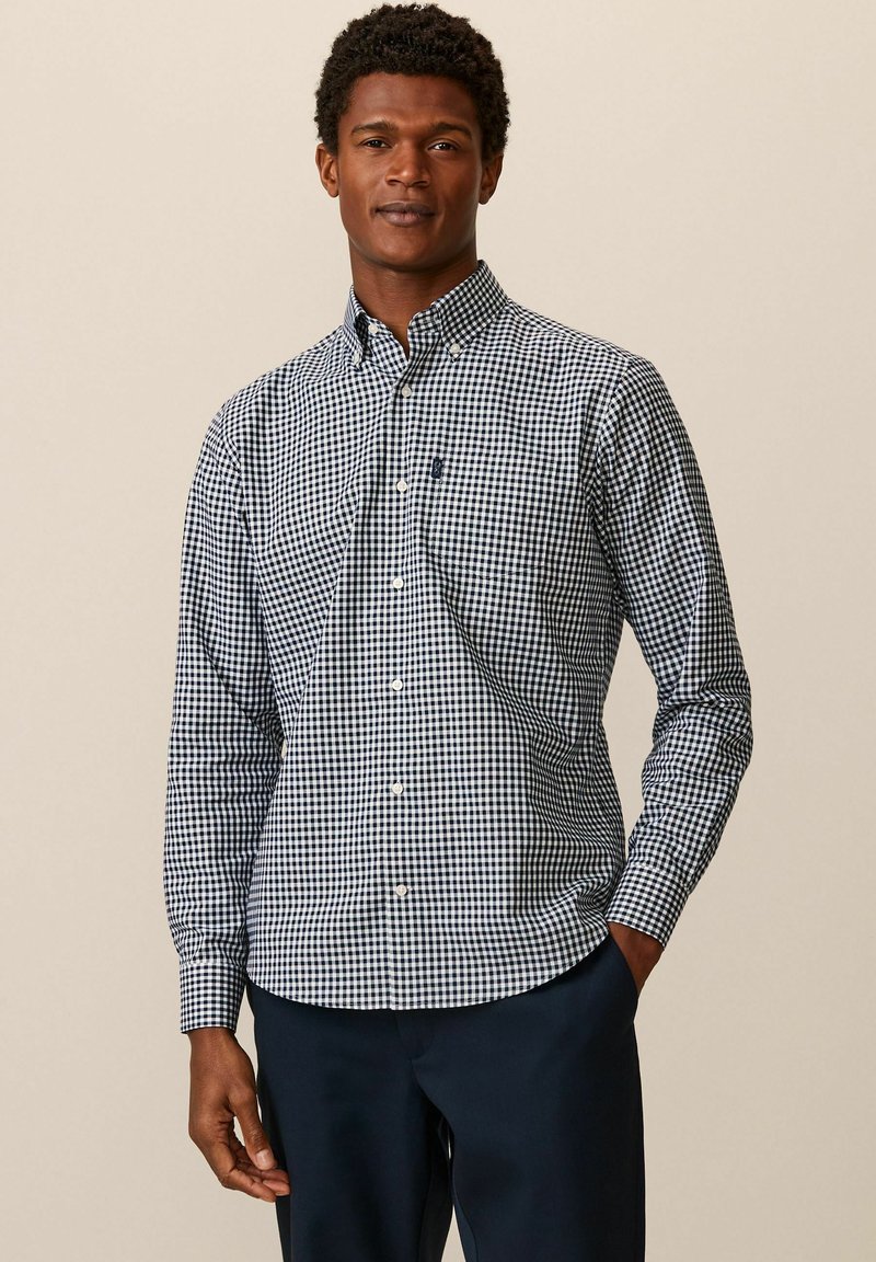 Chemise à carreaux bleue et blanche, à manches longues, avec col, en tissu doux et avec une poche poitrine. Associée à un pantalon noir.
