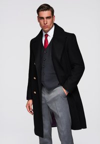 Manteau en laine noir avec de grands boutons dorés, associé à un gilet sombre et un pantalon gris clair. Texture lisse ; design classique et ajusté.