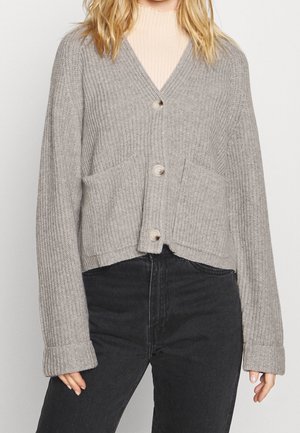 Kvinde iført en lysegrå ribstrikket cardigan med tre knapper, store lommer og vide ærmer over en beige top og sorte jeans.