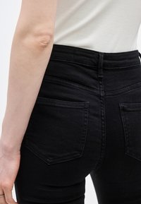 Κοντινή άποψη ενός ατόμου που φορά ψηλόμεσα μαύρα τζιν και λευκό τοπ, με ορατή την πίσω τσέπη και μέρος του χεριού.