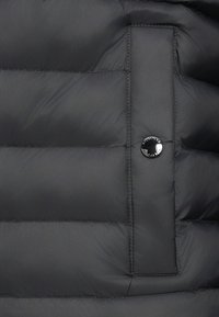 Svart quiltet stoff med horisontal søm og en vertikal lommeklaff sikret med en rund svart trykknapp.