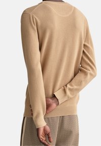 Pull en tricot beige avec tissu texturé, manches longues et poignets côtelés. Présente un col arrondi et une silhouette ajustée.