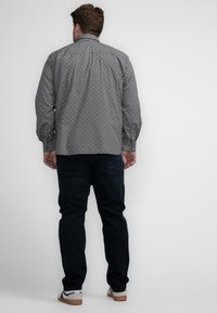 Man met kort haar in een grijs patronen lange mouw shirt, donkere jeans en witte sneakers staat met zijn gezicht naar een eenvoudige witte muur.