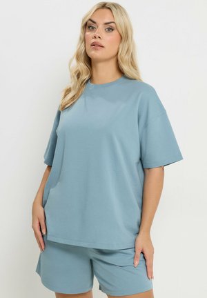 Blondynka w luźnej, jasnoniebieskiej, oversize koszulce i pasujących szortach, stojąca ze swobodnie opuszczonymi rękami.