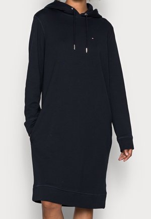 Kobieta nosząca luźną czarną sukienkę z kapturem z długimi rękawami, przednimi kieszeniami i małym logo Tommy Hilfiger na piersi.