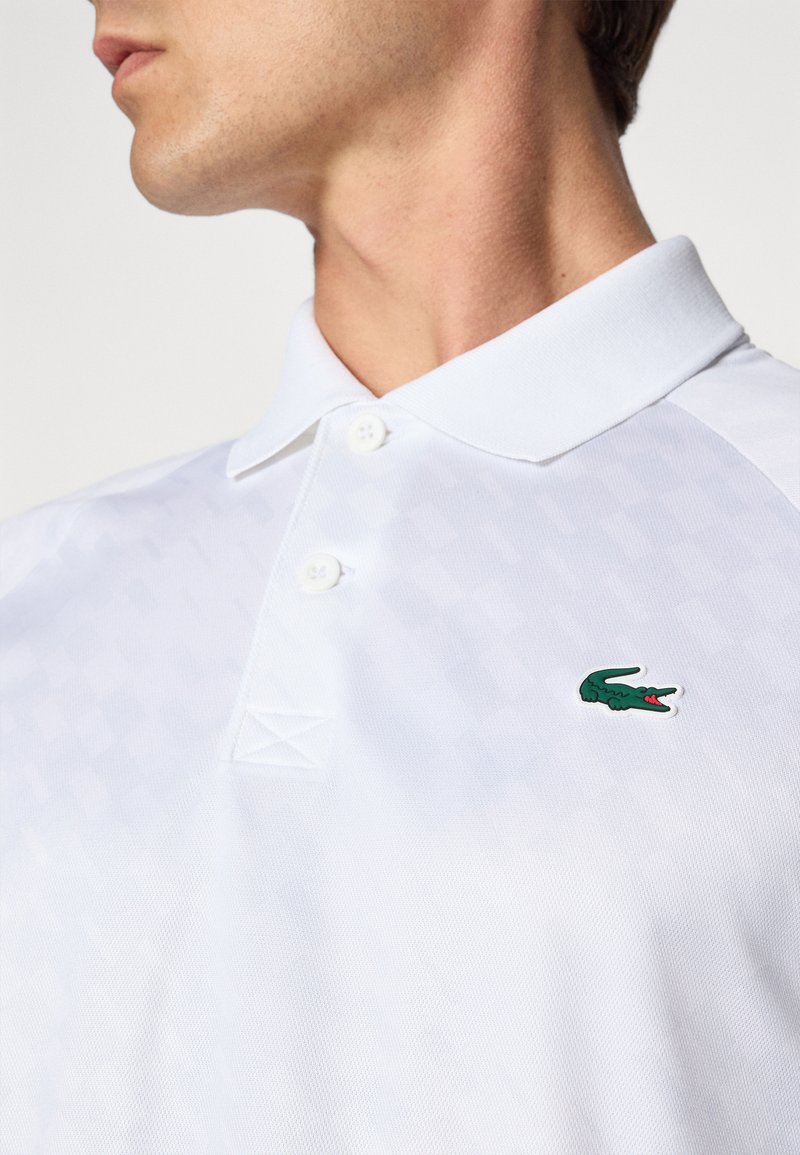 Polo blanc avec un motif texturé, deux boutons au col et un logo de crocodile brodé vert sur la poitrine gauche.