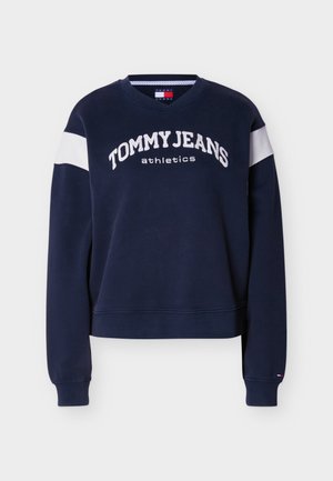 Tumši zils džemperis ar apaļu kaklu, baltu svītru uz piedurknēm un baltu uzrakstu "TOMMYJEANS athletics" uz krūtīm. Izgatavots no kokvilnas maisījuma materiāla.