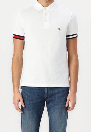 Mężczyzna w białej koszulce polo z mankietami w czerwono-czarno-białe paski i niebieskich dżinsach, stojący na tle białego tła.