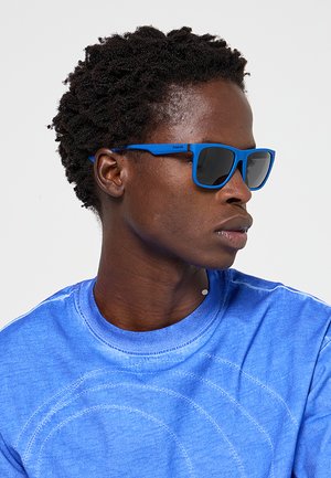 Uomo con capelli corti e ricci che indossa occhiali da sole blu e una camicia blu guarda di lato contro uno sfondo chiaro uniforme.