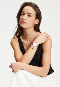Armbåndsur med hvid urskive, rosegold metalarmbånd og enkle timemarkører. Modellen bærer en sort ribbet tanktop og lagdelte armbånd.