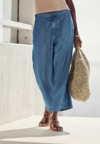 Lichtblauwe, loszittende broek van zacht materiaal, met een trekkoord in de taille en twee voorzakken. Hielkleurige sandaaltjes met bandjes en een geweven tas.
