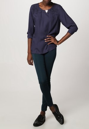 Blusa blu navy con scollatura decorativa, maniche a tre quarti, abbinata a pantaloni slim-fit verde scuro e scarpe nere texture.