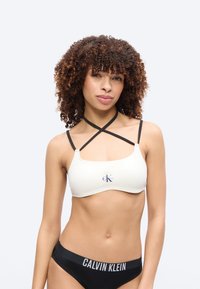 Γυναίκα με σγουρά μαλλιά που φοράει λευκό crop top με μαύρες διασταυρωμένες τιράντες και μαύρο εσώρουχο Calvin Klein, στέκεται μπροστά σε απλό φόντο.