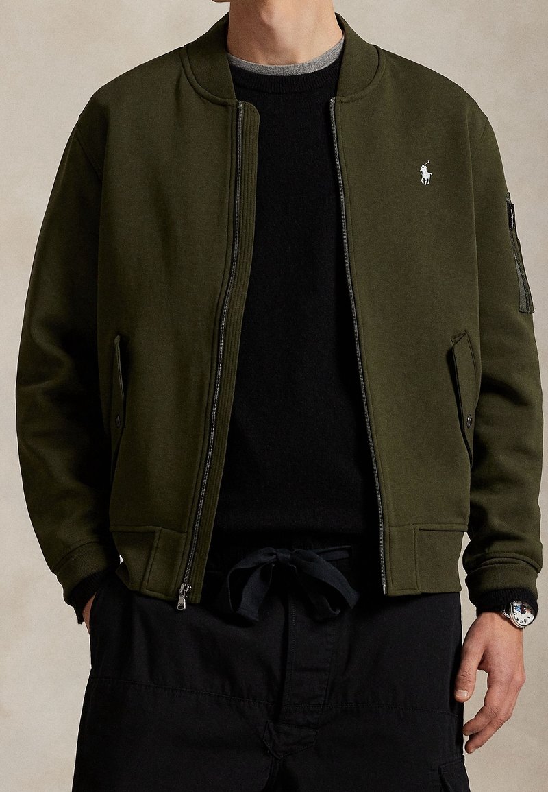 Homme portant une veste kaki ouverte avec un logo blanc sur la poitrine, un pantalon noir et une montre noire au poignet droit.