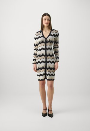 Strikkekjole med et chevron-mønster i beige, svart og krem, med V-hals og lange ermer, avsluttet med en bølget kant.