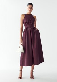 Burgundska, brezrokavna obleka s stisnjenim pasom in odprto krilo. Ima stranske žepke in je stilizirana z belo angularno torbico in črnimi petkami.