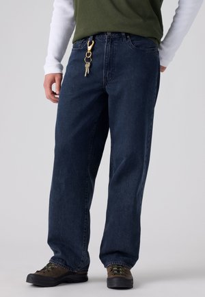 Personne portant un jean large bleu foncé, des chaussures marron, une chemise verte, un sous-pull blanc à manches longues, avec des clés accrochées à la boucle avant de la ceinture.
